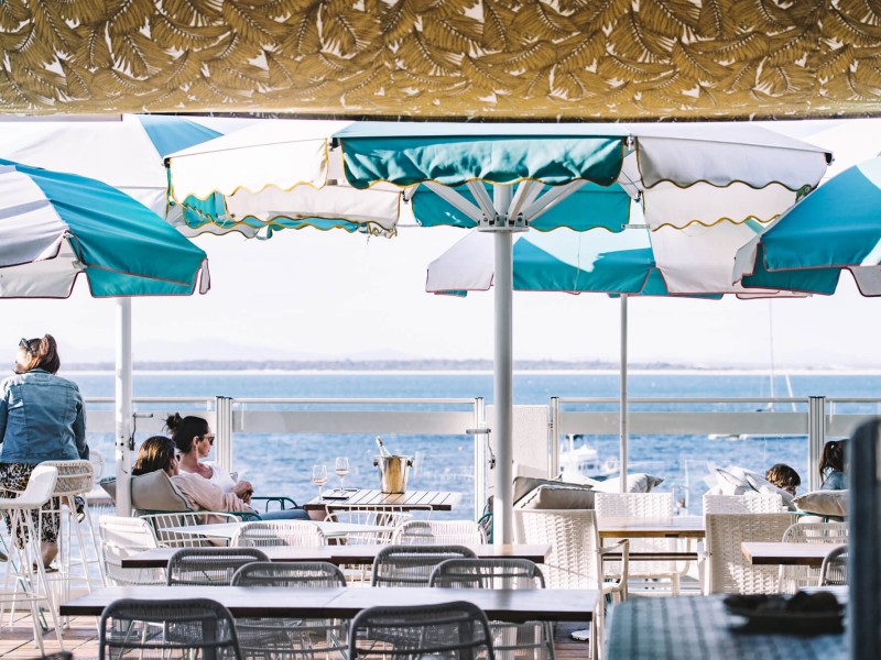 People relax at Shoal Bay Country Club under striped umbrellas. The scene conveys a calm, leisurely vibe.